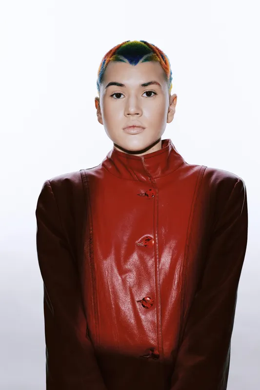 Serious woman with colorful short hair wearing a red leather jacket, standing confidently against a plain white background in a striking, modern portrait style.