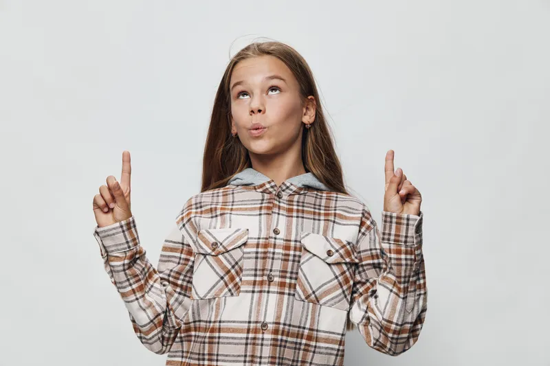 Girl in plaid shirt looks up with curiosity and points fingers upwards on both sides, standing against gray background with thoughtful expression and long hair.