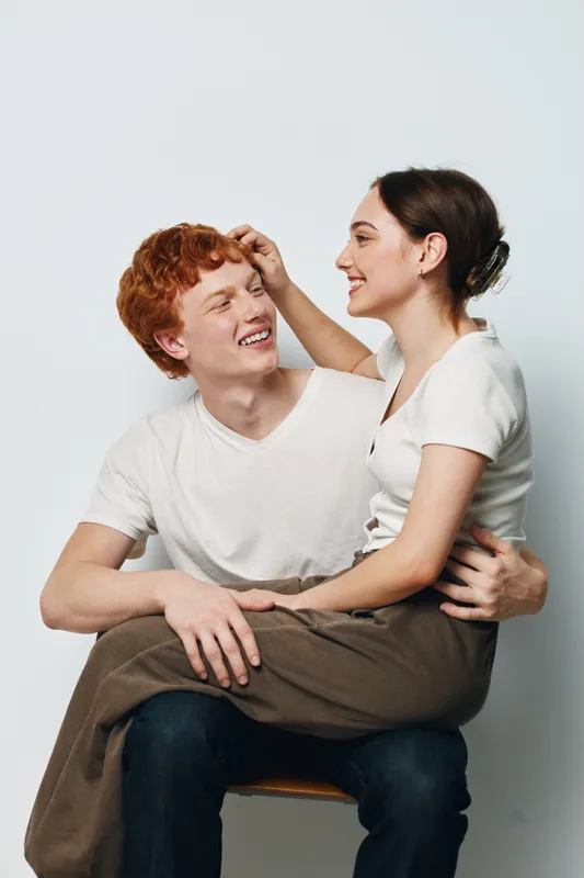 Young couple smiling and sitting close together in casual clothes on white background. Happy man and woman looking at each other with affection and joy in studio portrait.