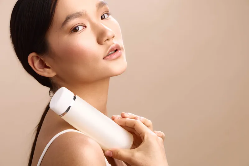 asian woman uses a sleek beauty device against her shoulder in soft studio light, exuding calm confidence and self care, a modern moment of personal wellness.