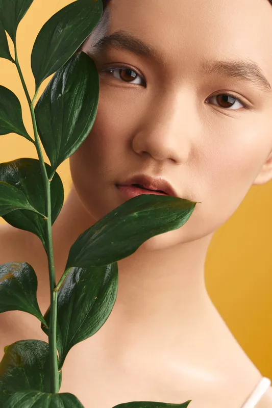 Asian woman is photographed with a leafy plant against a bold yellow background, exuding calm and natural elegance in a studio portrait, inviting fresh aesthetic vibes.