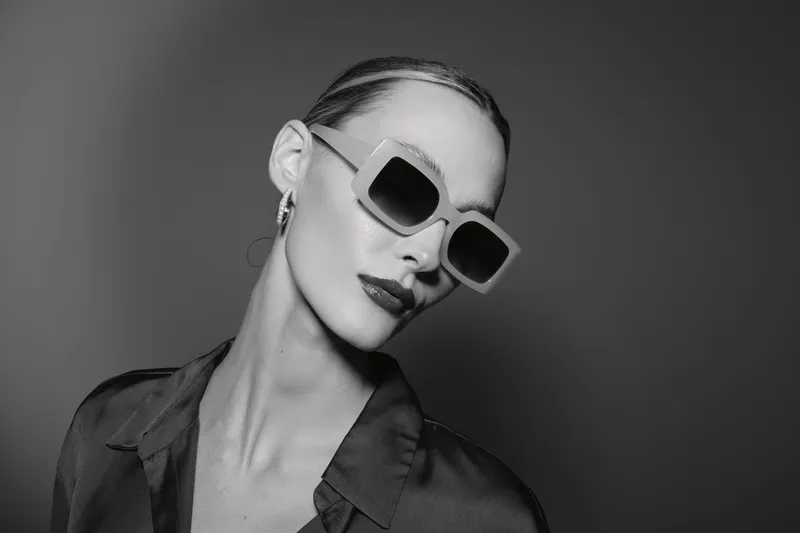 Stylish white woman with pale skin wearing large sunglasses and dark lipstick poses against grey background in fashionable black outfit.