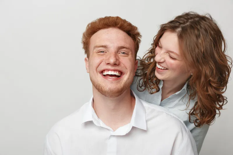 Happy young man and woman laughing together on white background. Casual clothing, joyful mood, friendship and positive emotions concept with natural light and studio setting.