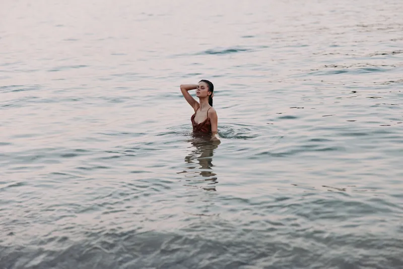 A lone woman stands waist-deep in calm sea water, hand at hair, soft light glinting on gentle ripples, creating a serene, intimate mood of quiet summer reflection.