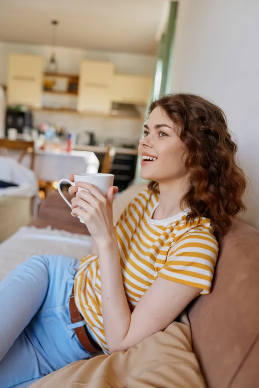 A woman enjoys a relaxed moment at home, resting on a sofa, holding a mug and smiling. A cozy, casual lifestyle scene emphasizing relaxation, comfort, and everyday wellbeing.