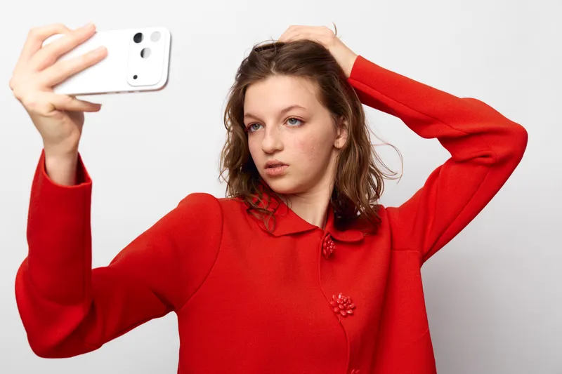 Young woman in red sweater takes a selfie with smartphone, posing with hand on head against white background. Concept of social media, self-portrait, and casual fashion.