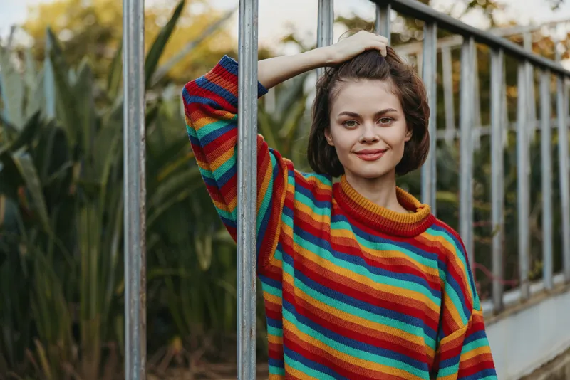 Young woman in her twenties wearing a colorful striped sweater, smiling and leaning on a metal railing in an urban garden with short hair, casual outfit and relaxed lifestyle portrait.