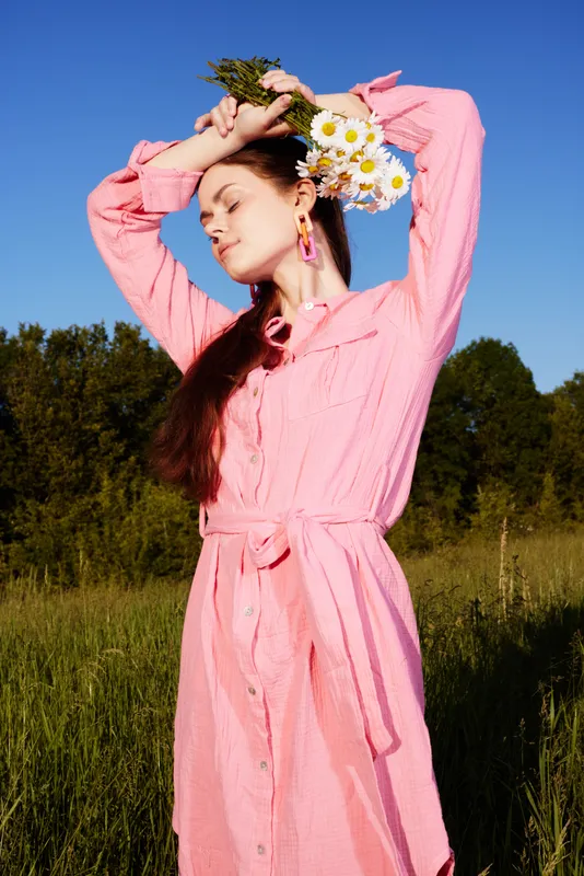 Stylish young woman in a pink dress with flowers, joyful expression, standing in a green field under a clear blue sky, capturing the essence of summer