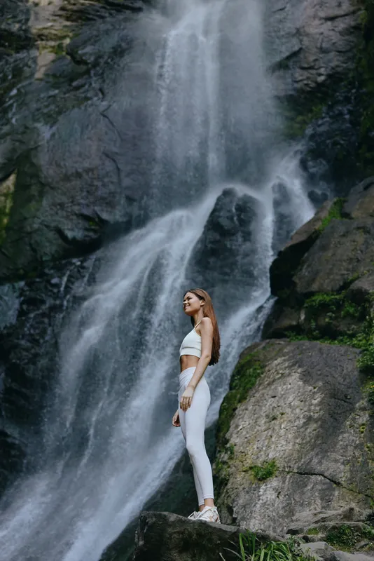 woman standing near a waterfall wearing white activewear and sneakers, surrounded by rocky cliffs and lush greenery in a natural outdoor setting
