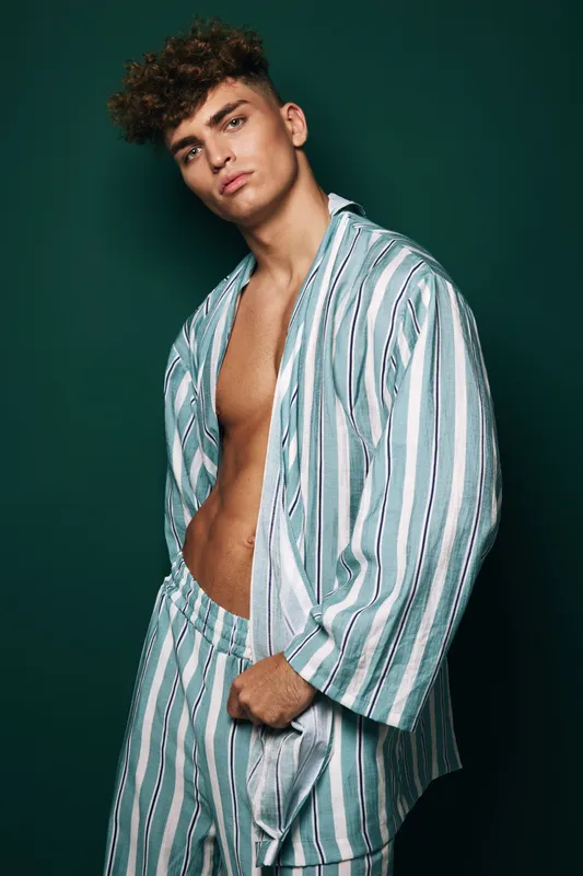 Young man with curly hair wearing striped pajamas posing against dark background. Casual fashion, relaxed style, male model with confident expression and open shirt.