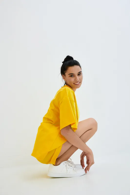 young woman crouching in yellow dress and white sneakers, smiling confidently against plain white background, casual style and modern fashion