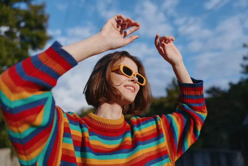 Woman smiling in colorful striped sweater and yellow sunglasses, short hair bob, outdoor portrait under sunny blue sky, joyful pose and casual fashion travel lifestyle.