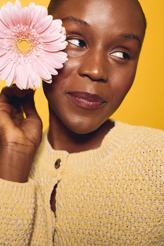 Close-up of a smiling black woman holding a pink flower near her face against yellow background. Portrait of natural beauty, dark skin, and floral concept with warm lighting and cozy sweater.