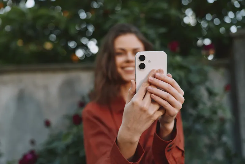 Smiling woman in a brown jacket takes a selfie outdoors, capturing a casual lifestyle moment with a garden backdrop and warm, natural light