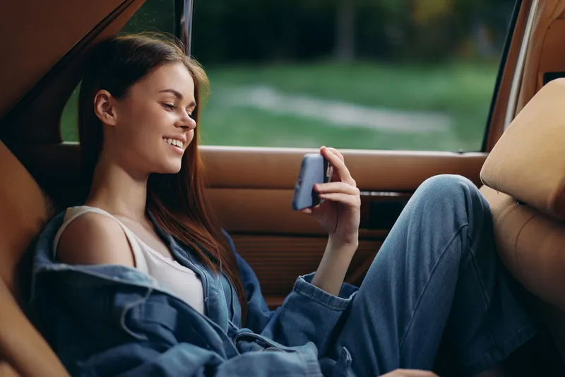 Happy young woman in casual denim outfit enjoying her smartphone in a car, expressing relaxation and joy in a natural outdoor setting.