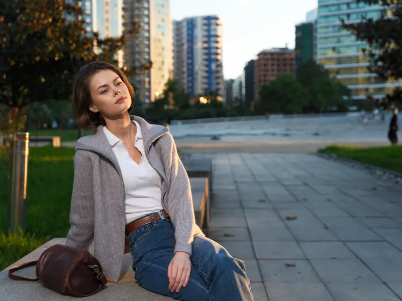 A calm professional woman sits on a bench in an urban park, wearing a light sweater and jeans. She gazes into the distance with poise, focus, and quiet confidence.