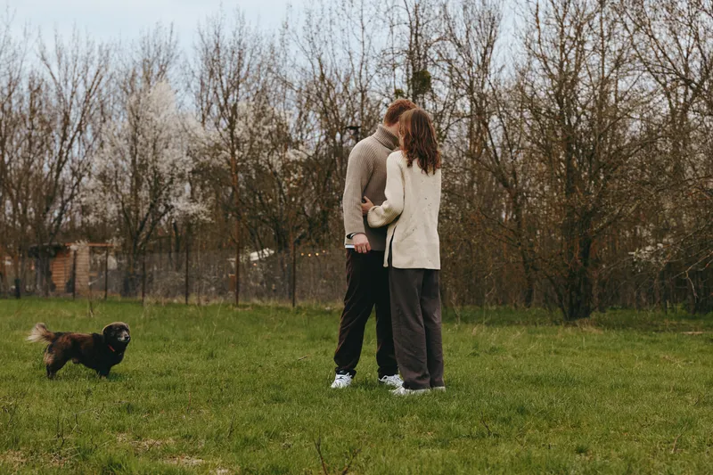 Couple sharing a gentle kiss in a sunlit park, holding hands, a small dog nearby, casual jackets and jeans, open grass and trees in the background, candid lifestyle moment, warm, intimate atmosphere,