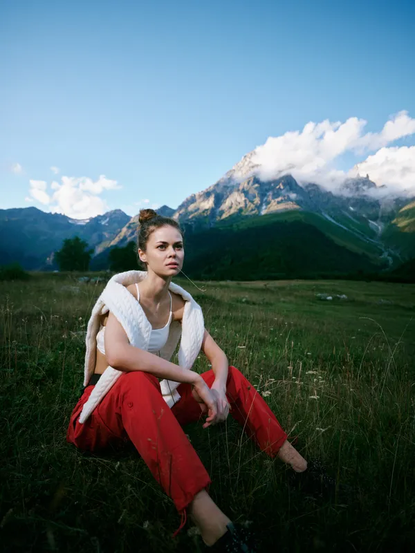 Woman sitting in a meadow with red pants and white shawl, mountains in the background, blue sky and clouds, a serene outdoor fashion moment with natural light