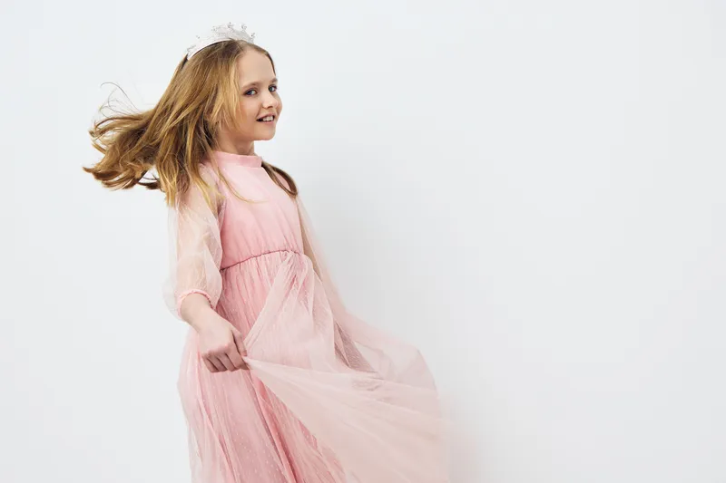 Joyful young girl in a light pink dress twirling with flowing fabric against a soft white background, exuding happiness and innocence in a playful moment