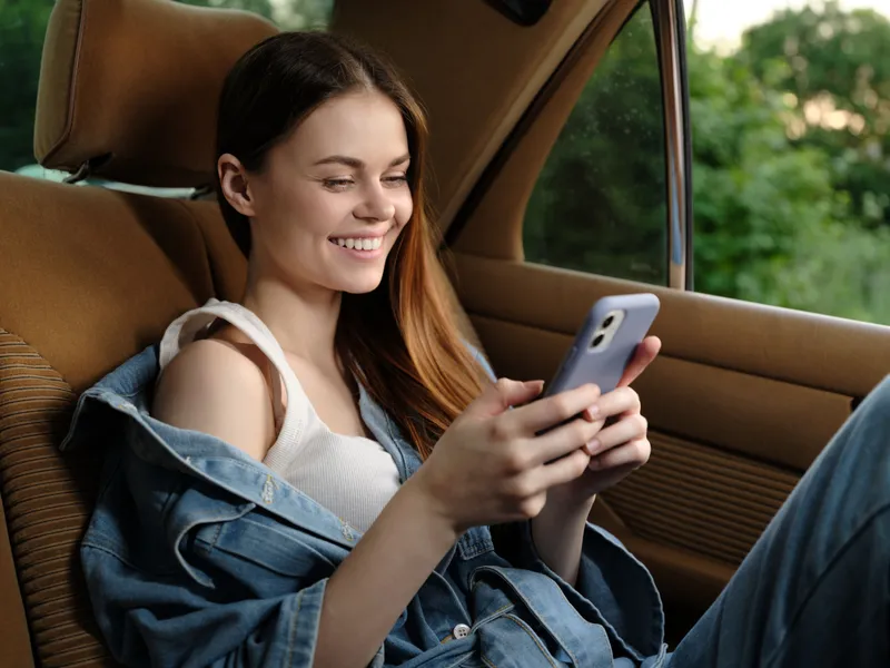 Young woman enjoying her smartphone in a car during a summer day, showcasing happiness and casual style with a denim jacket.