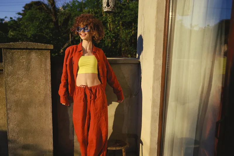 Smiling woman with curly hair wearing a bright yellow crop top and orange suit, standing outdoors. Casual lifestyle portrait conveys confidence, sunny mood, and vibrant summer style