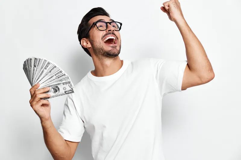 Young smiling man holding cash dollars cheerful and excited celebrating success with arms raised isolated on plain solid light background. Concept of happiness