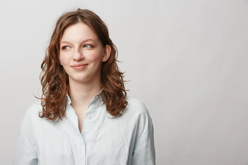 Young woman with curly brown hair wearing white shirt looks sideways with slight smile on light background. Concept of casual style, natural beauty, and positive mood.