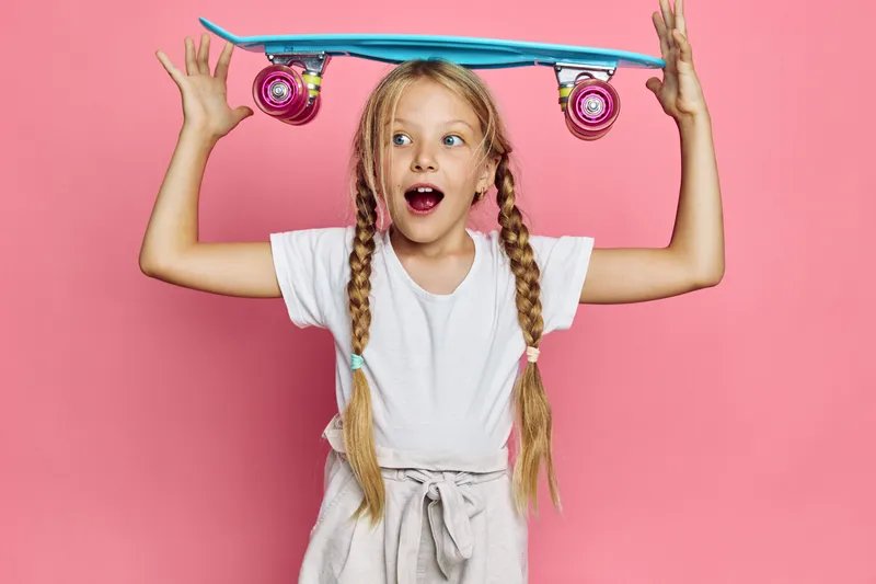 Happy girl with blonde braids holding a blue skateboard above her head on a pink background. Studio shot with surprised expression.