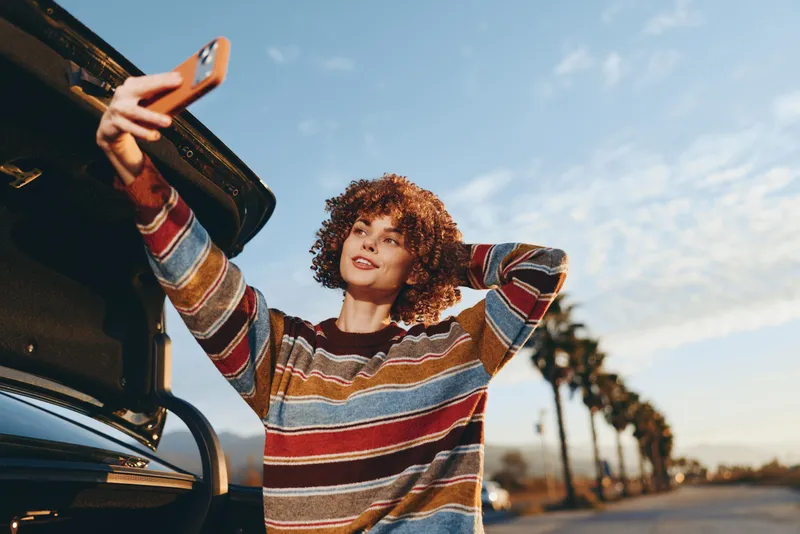 Woman car phone striped sweater curly hair sunset lifestyle outdoors, young woman in striped sweater holds smartphone beside car at sunset, sunlit palm lined road, casual lifestyle smile