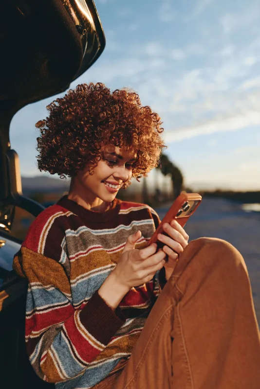 Smiling woman lifestyle portrait in a car at sunset, curly hair framing her face as she reads a smartphone screen, relaxed travel moment, casual sweater, warm light outside today.