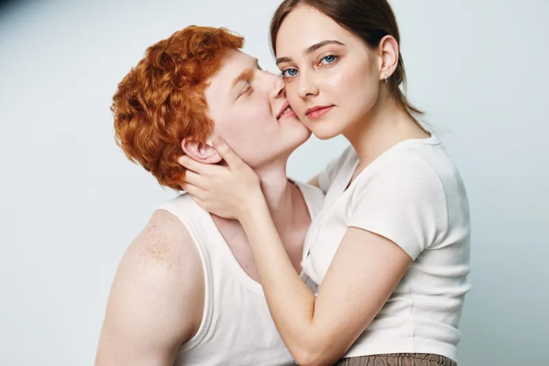 Couple with red hair and pale skin embraces each other, showing affection and tenderness in casual white tops with soft expressions on plain background.