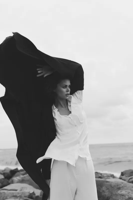 Woman on a windy beach wearing a flowing white dress as a dark cape billows, creating dramatic monochrome motion by the ocean horizon