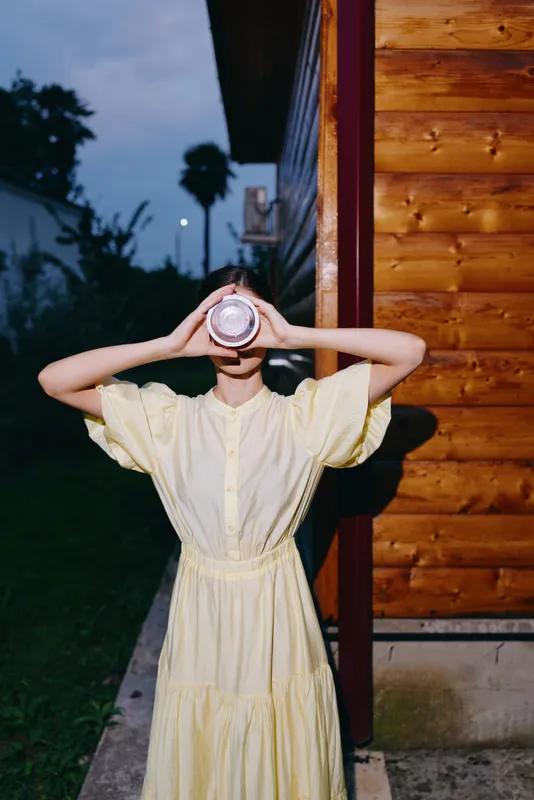 Portrait of a woman in a yellow dress standing outdoors by a wooden building, holding a round object to her face, creating a playful mysterious mood at dusk.