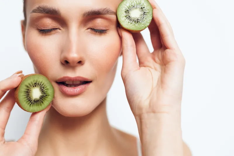 A closeup of a woman with clear skin holding two kiwi halves near her face with closed eyes. Concept for natural beauty, skincare, and healthy lifestyle.