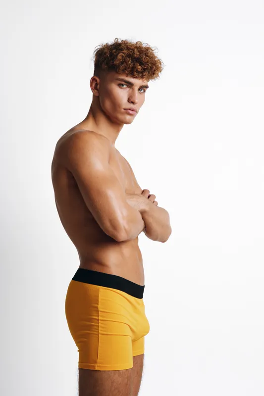 Young man with curly hair posing shirtless in yellow boxer briefs against white background. Side view of fit male model with arms crossed and serious expression in studio lighting.