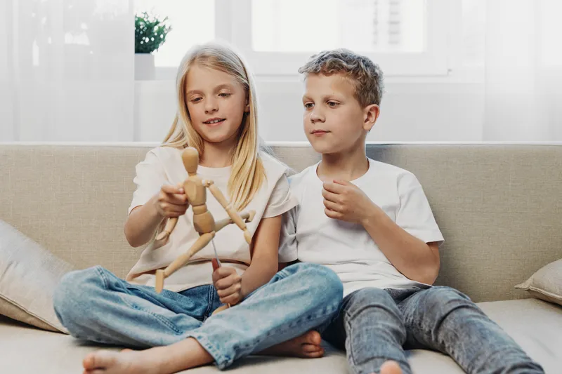 Two children sitting on sofa indoors playing with wooden mannequin toy, casual clothes, natural light, home environment, childhood and creativity concept.