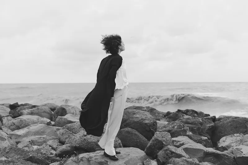Woman stands on rugged rocks by the sea, coat billowing in the wind, facing the horizon; monochrome mood captures solitude, movement, and coastal atmosphere