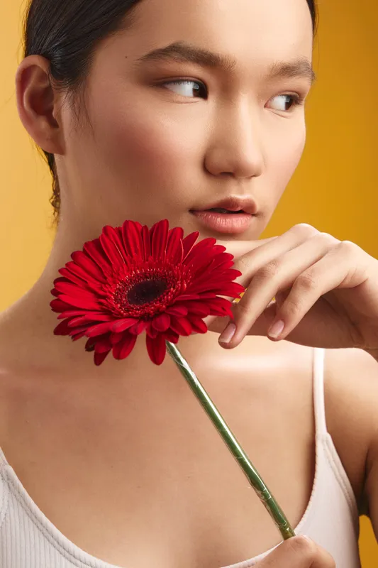 Asian woman gazes at a vibrant red flower up close, her calm expression and soft studio lighting creating a serene, intimate portrait with a delicate floral motif.