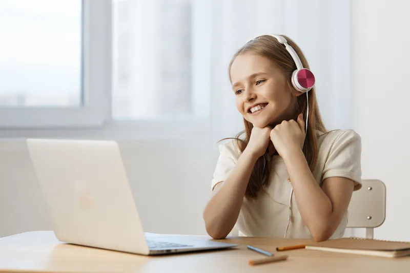 Young girl with headphones smiling at a laptop in a bright room, expressing joy and excitement while learning or playing online, perfect for educational content