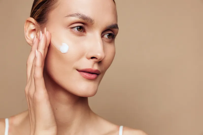 Closeup of young woman applying moisturizing cream on face with fingers, smooth skin, natural makeup, neutral background, skincare routine, soft lighting, beauty concept.