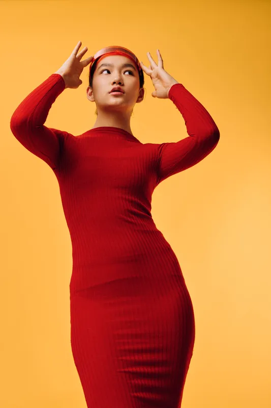 Woman wearing red dress and headband posing with hands near head against yellow background. Stylish female model in long slim dress expressing thoughtful emotion in studio.