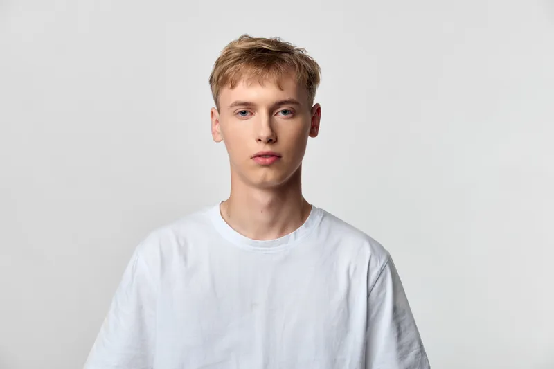 Young man in a white t shirt stands before a neutral background in a studio shot, presenting a calm expression and modern simplicity with clean lighting and minimal styling, short blond hair.