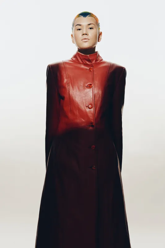 Woman with short hair wears a long red leather coat standing against a neutral background, featuring modern fashion and confident posture in stylish outerwear.