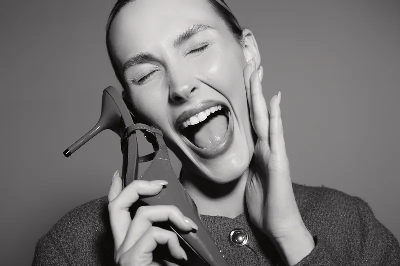 Happy Caucasian woman with fair skin holding high heel shoe near face with eyes closed and mouth open. Concept of joy, fashion, and playful mood in studio black and white photo.