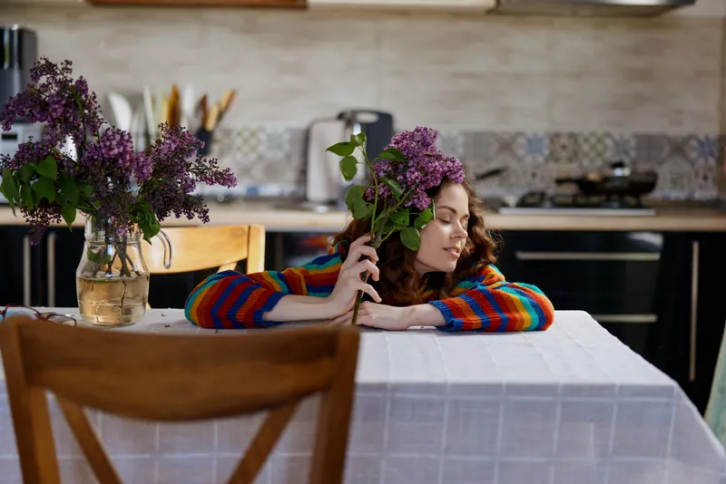 Young woman sits at a kitchen table with a bouquet, resting her cheek against flowers, enjoying a peaceful home moment in colorful sweater sleeves and soft natural light, mood warm and reflective.