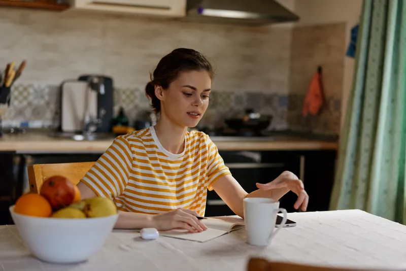 Cheerful woman indoors at a sunlit kitchen table, wearing a striped shirt, writing in a notebook, a mug nearby, capturing a calm domestic morning lifestyle