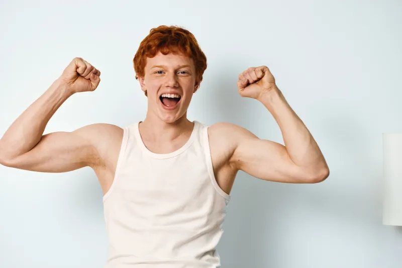 Young man with red hair and white tank top flexing muscles with excitement in bright room. Emotions, strength and energy shown through facial expression and fists up with smile.