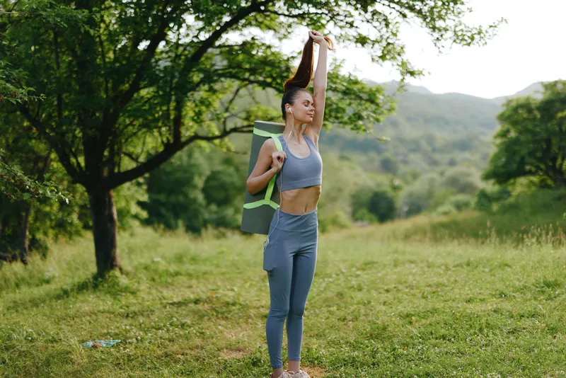 Fit young woman stretching outdoors with yoga mat, wearing athletic clothing in a serene natural setting, exuding confidence and wellness during summer