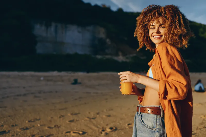 Woman curly hair beach smiling summer drink sunset outdoors lifestyle portrait, holding a glass of orange juice on a sandy shore, golden sunlight, warm glow, relaxed vacation mood, soft blur