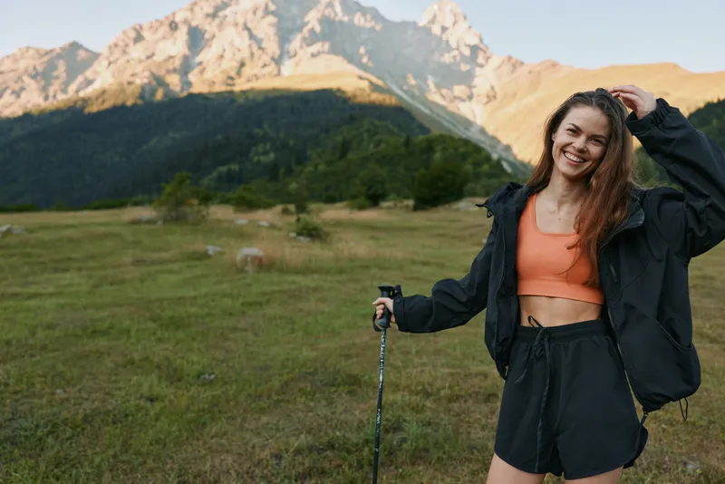 A cheerful young woman hikes in a mountainous landscape. She wears an orange crop top, black jacket and shorts, smiles at the camera, and rests on trekking poles, creating an energetic outdoor scene.